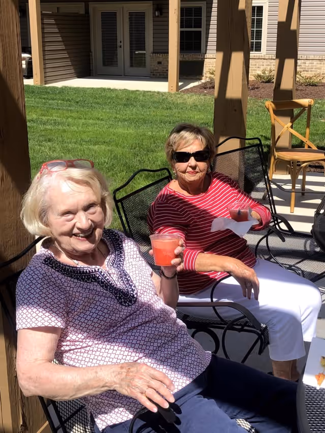 Two elderly women sitting outside on metal chairs under a wooden pergola. One woman is wearing a patterned short-sleeve shirt and holding a red drink, smiling at the camera. The other woman is wearing a red and white striped long-sleeve shirt, white pants, and sunglasses, also holding a red drink with a napkin. Behind them is a green lawn and a building with beige siding and windows.