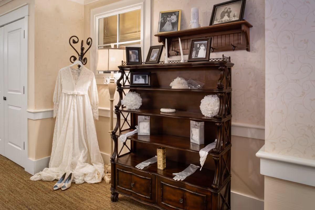 A vintage white wedding dress hanging on a decorative coat rack next to a floor lamp and a dark wooden shelf displaying framed black and white wedding photos, white gloves, and other wedding memorabilia in a softly lit room with patterned wallpaper.