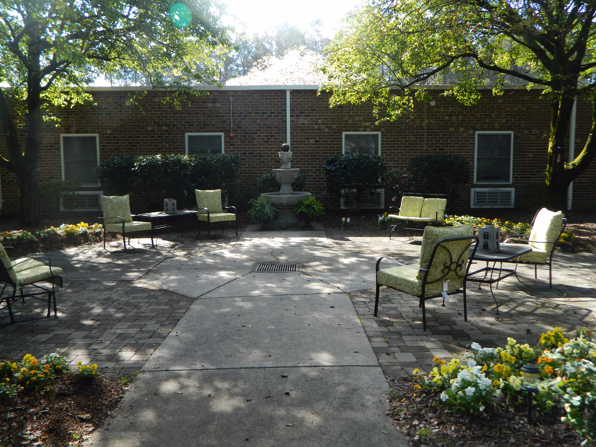Outdoor patio area with several cushioned chairs and small tables arranged around a central concrete space. There is a three-tiered stone fountain against a brick building wall, surrounded by bushes and flower beds. Trees provide partial shade over the area.