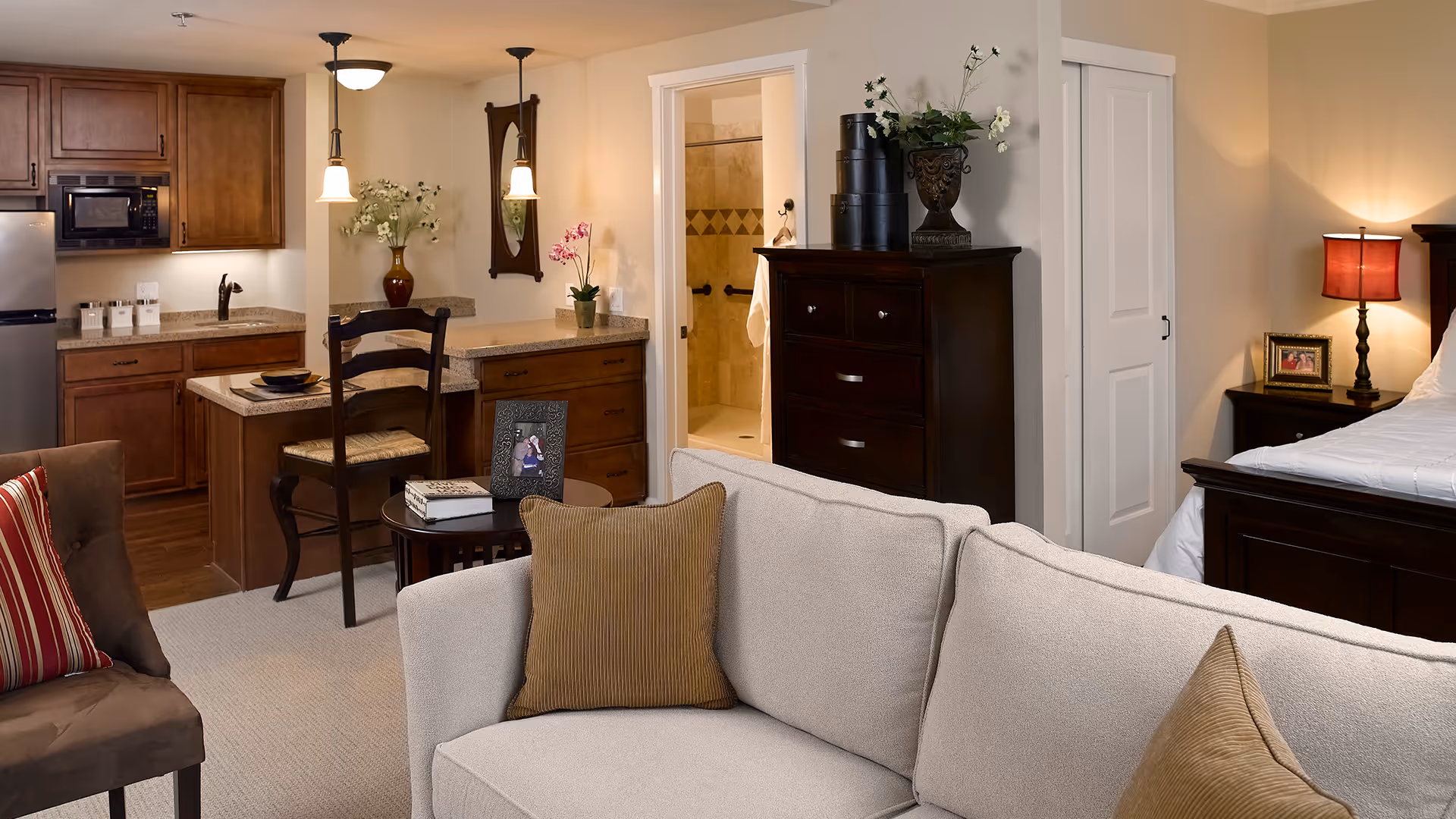A cozy studio apartment interior at Atria Golden Creek featuring a beige sofa with brown pillows in the foreground, a dark wooden chair and small round table with a photo frame and book, a kitchenette with wooden cabinets, a microwave, and a refrigerator. In the background, there is a bathroom with a walk-in shower, and a bedroom area with a dark wooden bed frame, nightstand, and a red lamp.