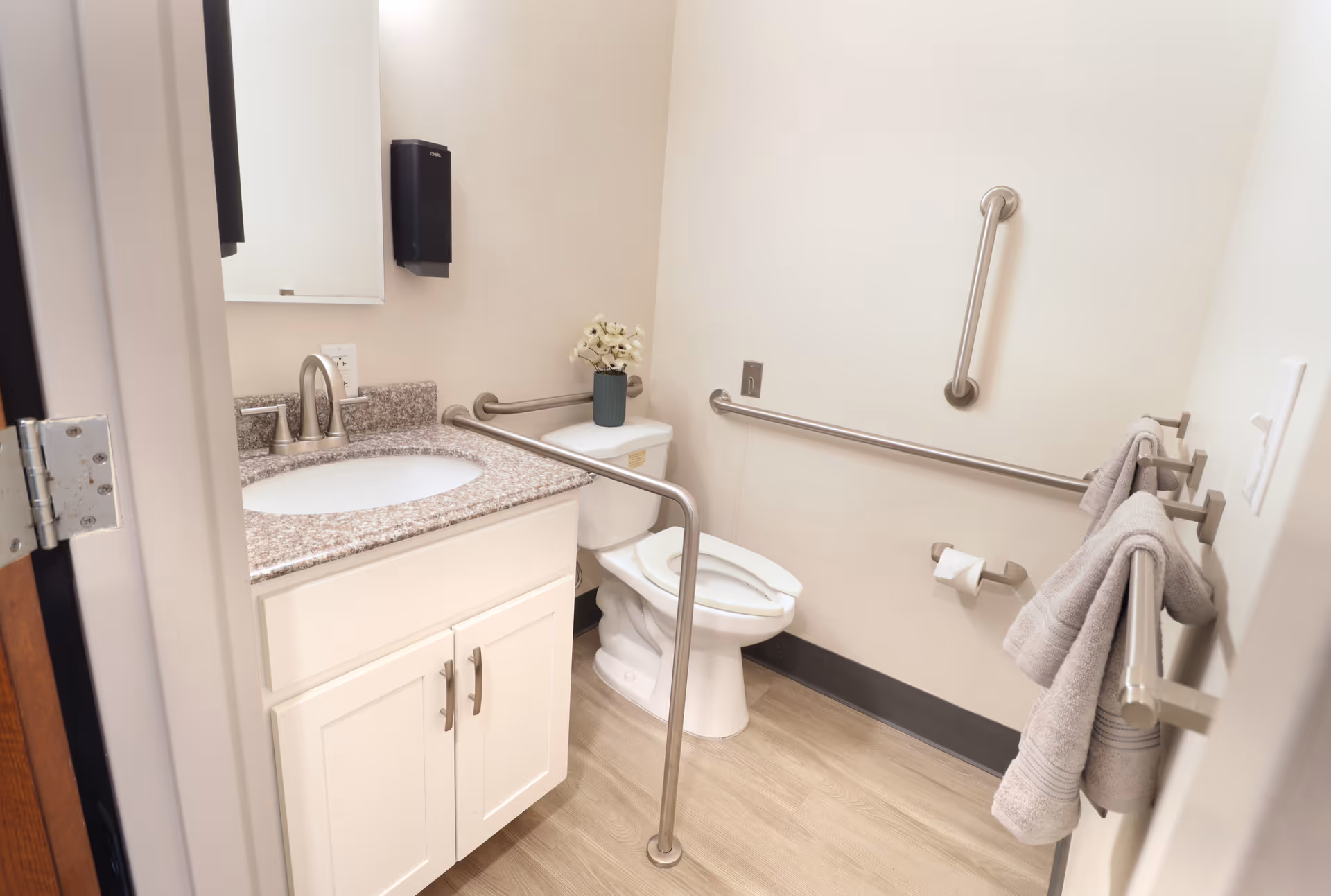 A clean and accessible bathroom with a white toilet, a white vanity with a granite countertop and sink, grab bars on the walls and around the toilet, a wall-mounted paper towel dispenser, a mirror above the sink, and two gray towels hanging on a towel rack.