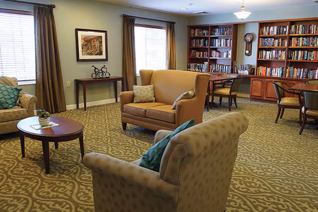 A cozy living room area with patterned carpet, two beige armchairs with green cushions, a beige loveseat with two pillows, a round wooden coffee table with a small plant, a wooden console table under two windows with brown curtains, and a large bookshelf filled with books along the back wall. There are also two wooden tables with chairs near the bookshelf.