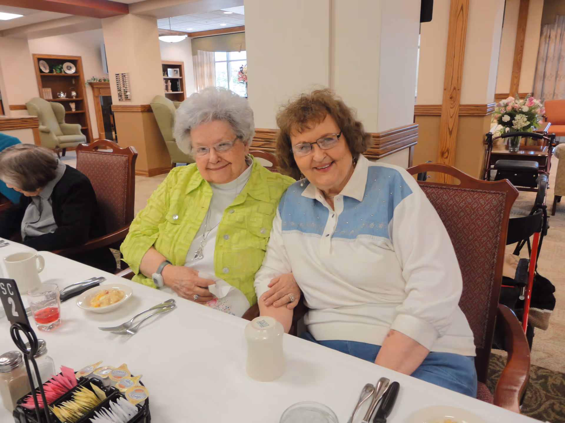 Two elderly women sitting closely together at a dining table in a senior living facility, smiling at the camera. The table is set with utensils, a small bowl of food, a glass with a red drink, and condiments. The background shows a cozy interior with chairs, shelves, and a floral arrangement.