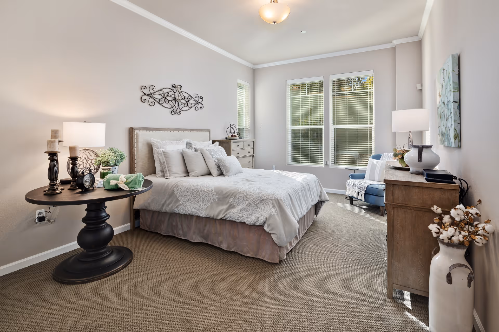 A bright and cozy bedroom with a large bed adorned with multiple pillows and a patterned bedspread. There is a round black side table with candles, a lamp, and decorative items on the left side of the bed. On the right side, there is a wooden dresser with a lamp, a vase, and other decor. A blue armchair with a throw blanket sits near the windows, which have white blinds letting in natural light. The walls are painted a soft beige color, and the floor is carpeted.