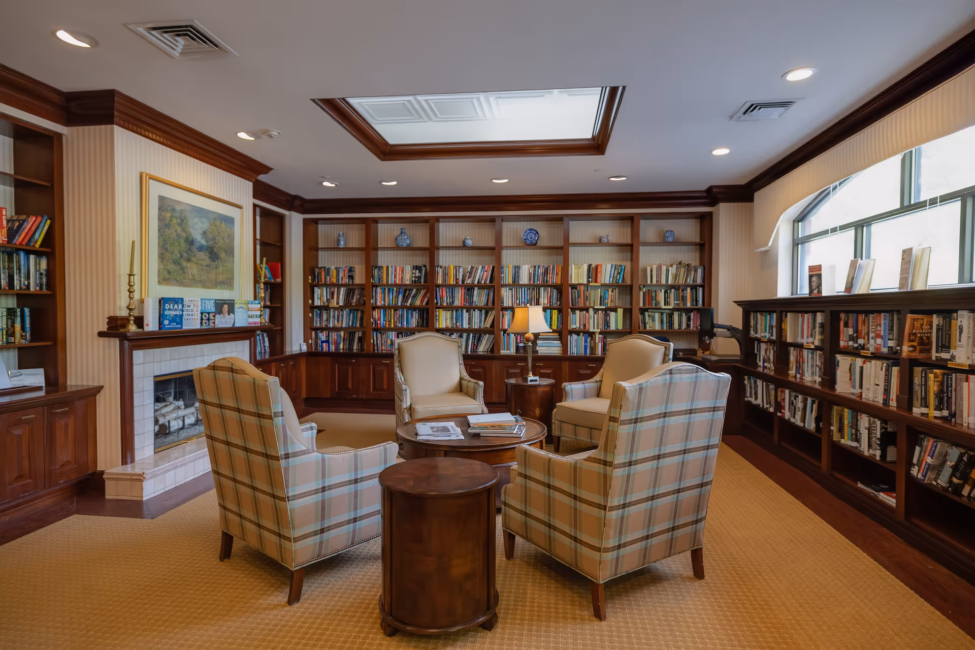 A cozy library room with wooden bookshelves filled with books lining the walls. Four upholstered armchairs, two in plaid and two in cream, are arranged around a circular wooden coffee table with books and magazines on it. There is a small round side table with a lamp between two of the chairs. A fireplace with a painting above it is visible on the left side. The room has a beige carpet and a skylight in the ceiling, with recessed lighting.