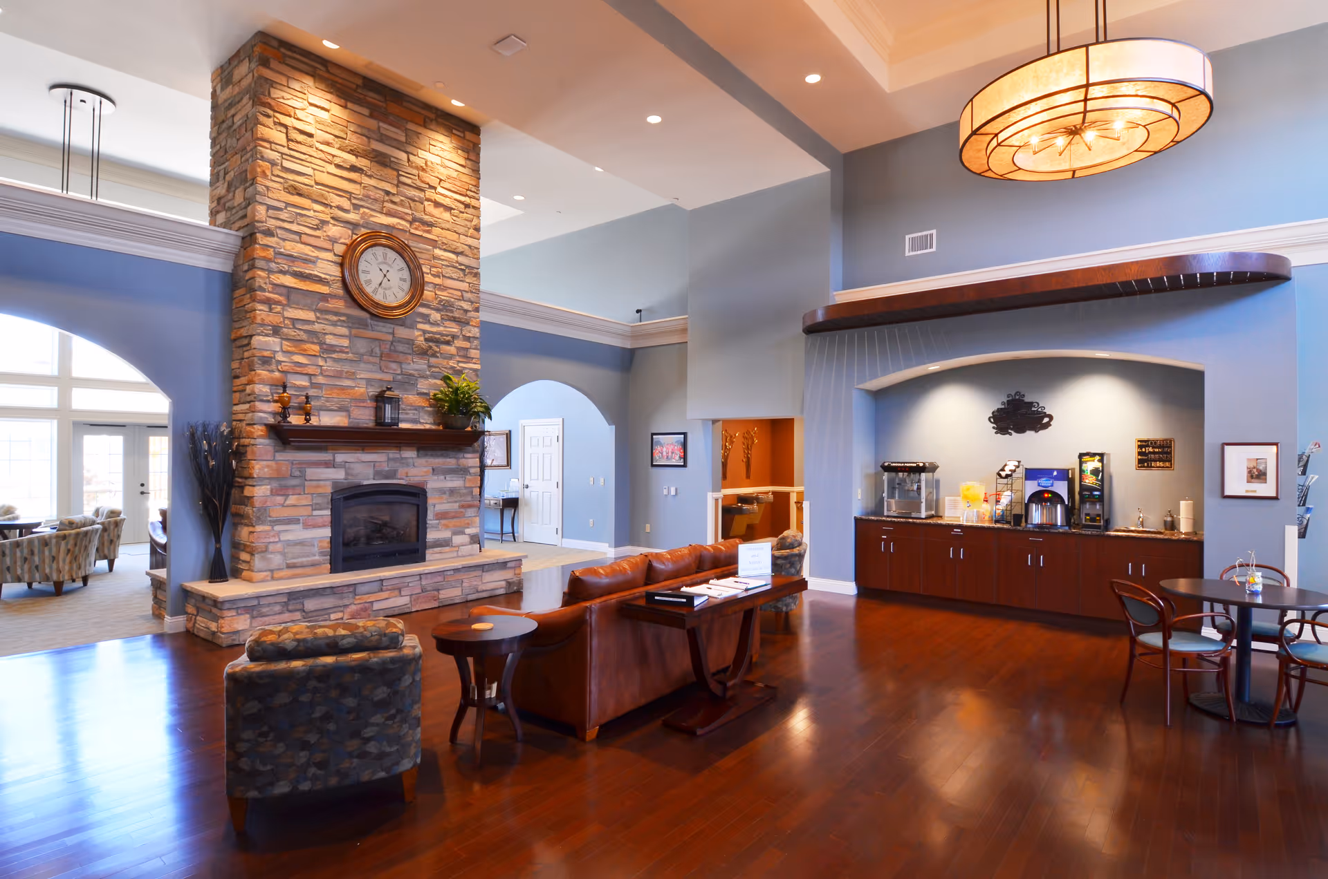 A spacious and well-lit common area in a senior living facility featuring a large stone fireplace with a clock above it, a brown leather sofa, a patterned armchair, a small round table, and a beverage station with coffee and juice dispensers under a curved alcove. The room has high ceilings, wooden flooring, and soft blue walls with white trim.