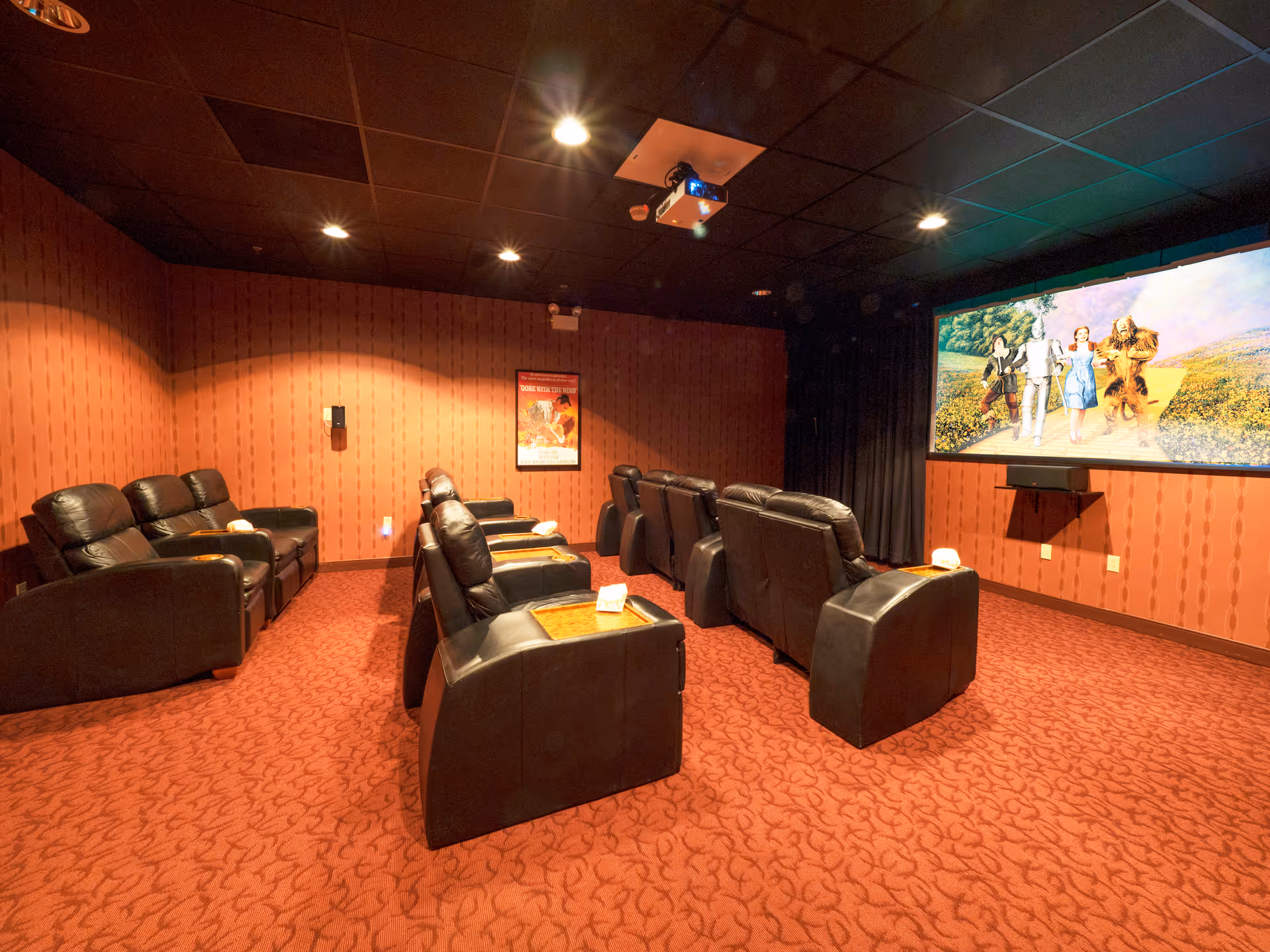 A cozy movie theater room with red patterned carpet and walls, black leather recliner chairs arranged in rows facing a large screen showing a scene from The Wizard of Oz. The ceiling is black with recessed lighting and a projector mounted in the center.
