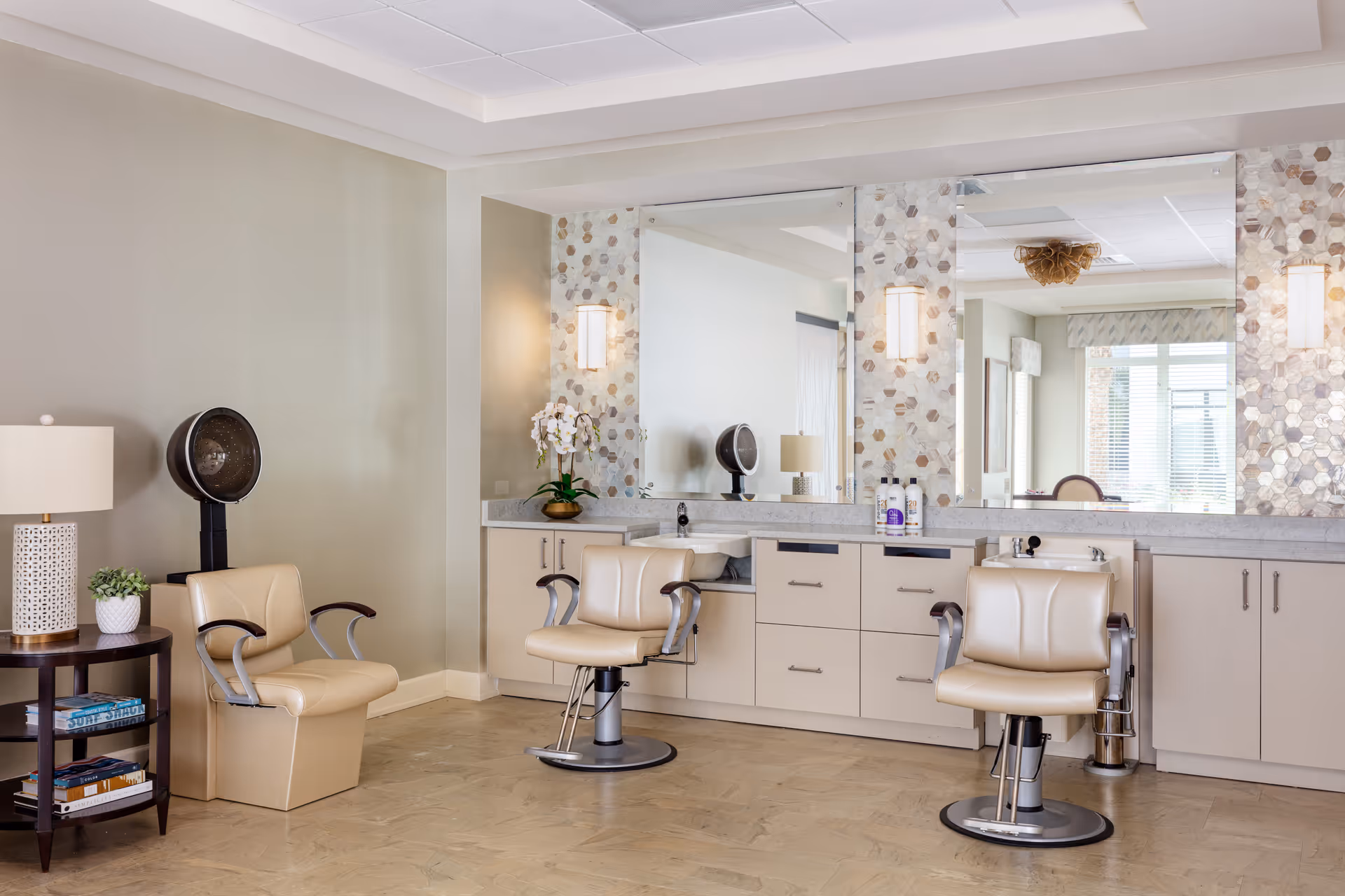 A bright and clean salon area with three beige salon chairs in front of a long counter with large mirrors. The counter has cabinets and drawers, with hair care products and a white orchid plant on top. There is a hair dryer station to the left and a small round table with a lamp and books beside it. The walls are light-colored with a decorative tile backsplash behind the mirrors.