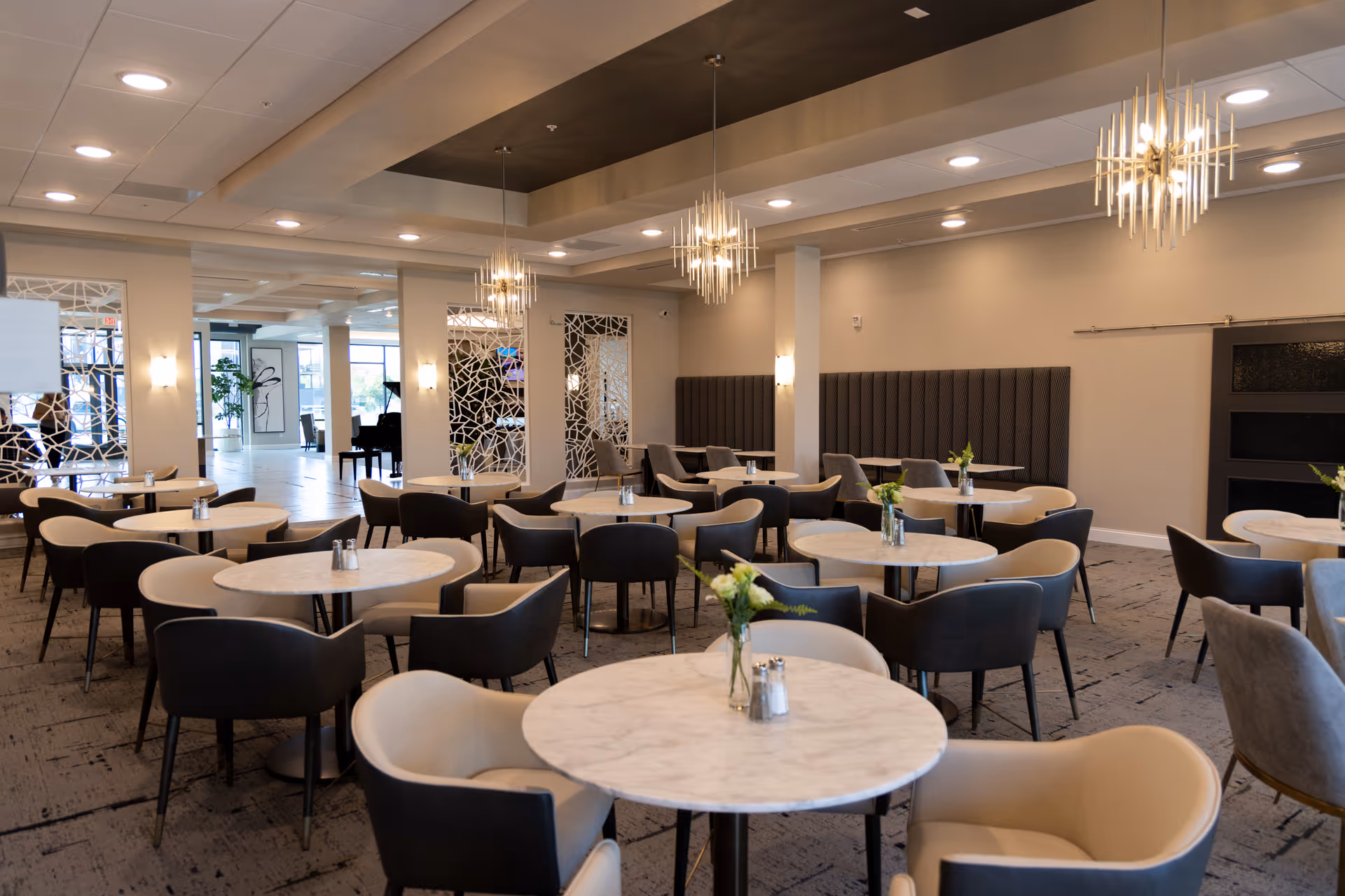 Spacious modern dining room with round marble tables, upholstered chairs, pendant lights, and small floral centerpieces.