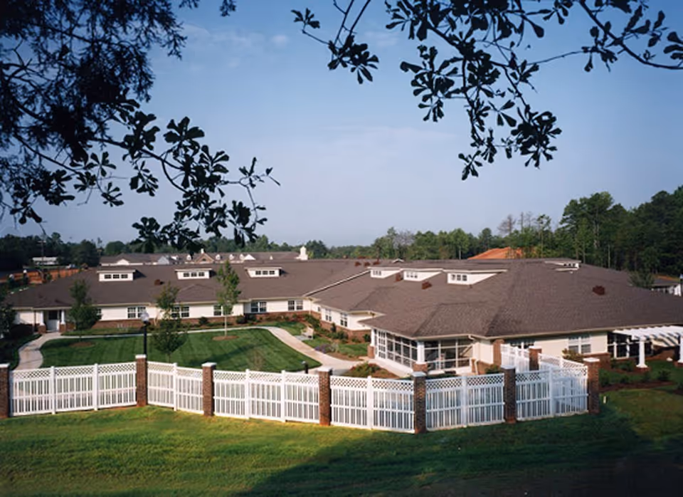 Exterior view of a single-story senior living facility with a large fenced-in grassy courtyard, surrounded by trees and a clear blue sky.