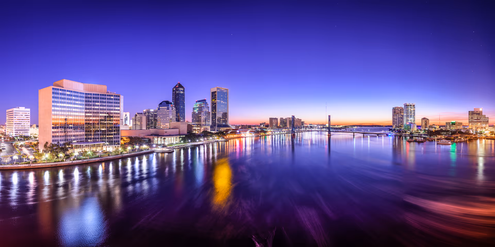 Panoramic view of a city skyline at dusk with buildings reflecting colorful lights on a wide river under a clear purple-blue sky.
