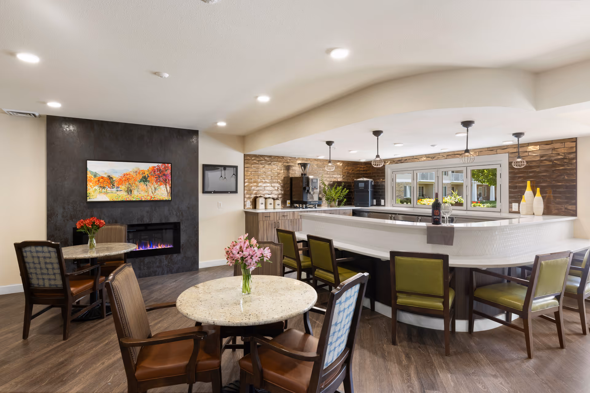 A cozy senior living common area with a modern fireplace set in a dark accent wall, a colorful landscape painting above it, and two round tables with chairs. The space features a large white countertop bar with green cushioned chairs, pendant lights hanging from the ceiling, and a window overlooking an outdoor area. The room has wood flooring and decorative vases on the counter.