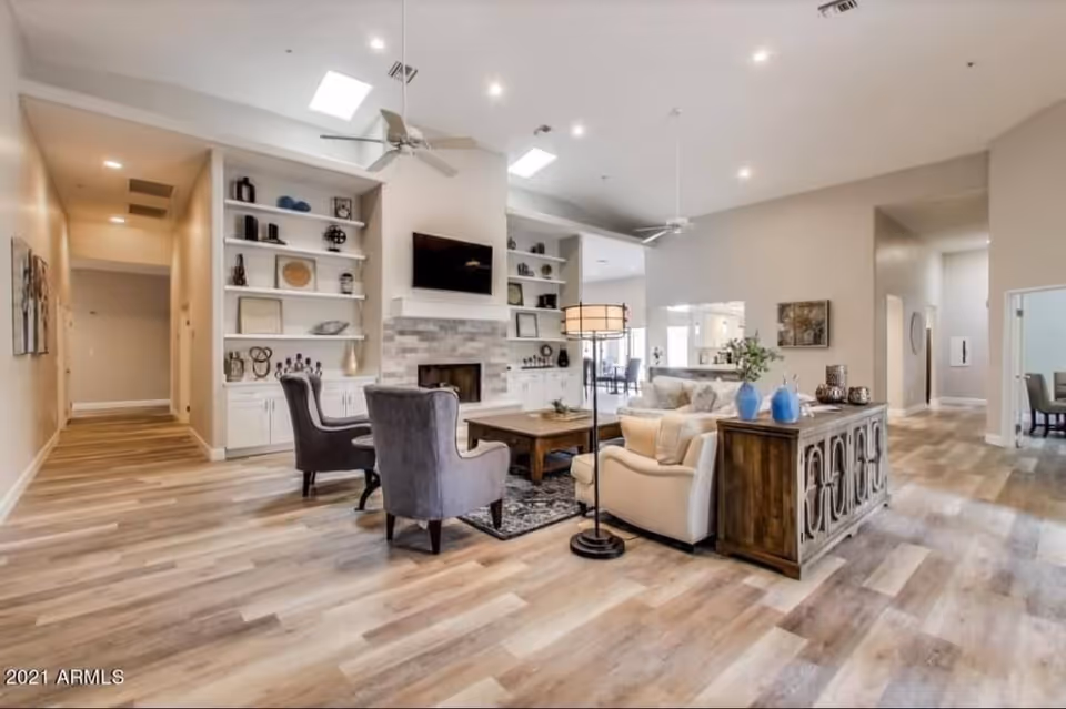 A spacious and well-lit living room area with high ceilings, ceiling fans, and recessed lighting. The room features a fireplace with a mounted TV above it, built-in shelves with decorative items, two gray armchairs, a beige sofa, a wooden coffee table, and a wooden sideboard with blue vases. The flooring is light wood, and there are hallways visible leading to other parts of the facility.