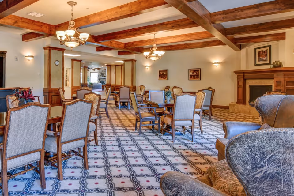 Spacious senior living dining room with multiple tables and chairs, wooden ceiling beams, chandeliers, and a fireplace.