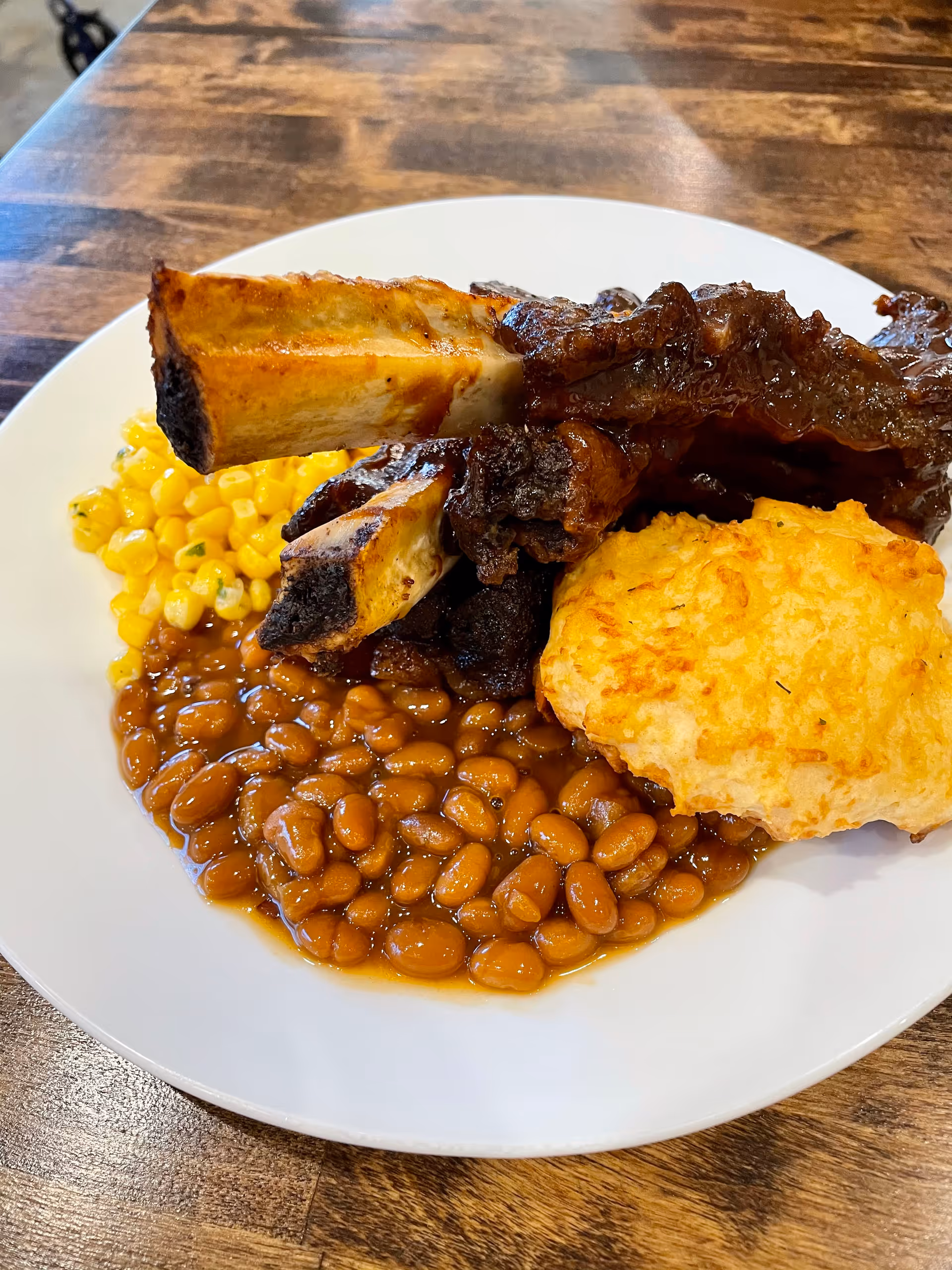 A white plate with a serving of baked beans, corn, a biscuit, and several ribs with a charred exterior, placed on a wooden table.
