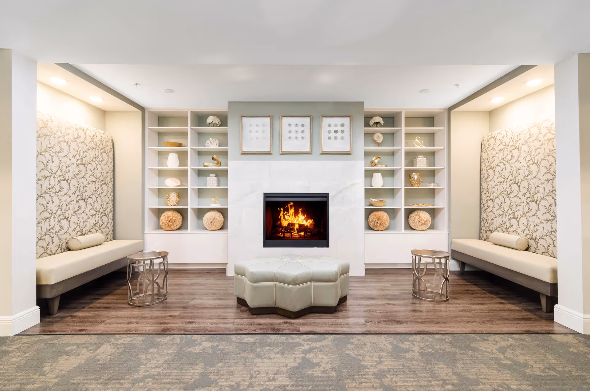 A cozy interior lounge area featuring a modern fireplace centered between two built-in shelves decorated with various ornaments. On either side of the fireplace are cushioned benches with patterned backrests and cylindrical pillows, accompanied by small round metallic side tables. The floor is a combination of wood and carpet.