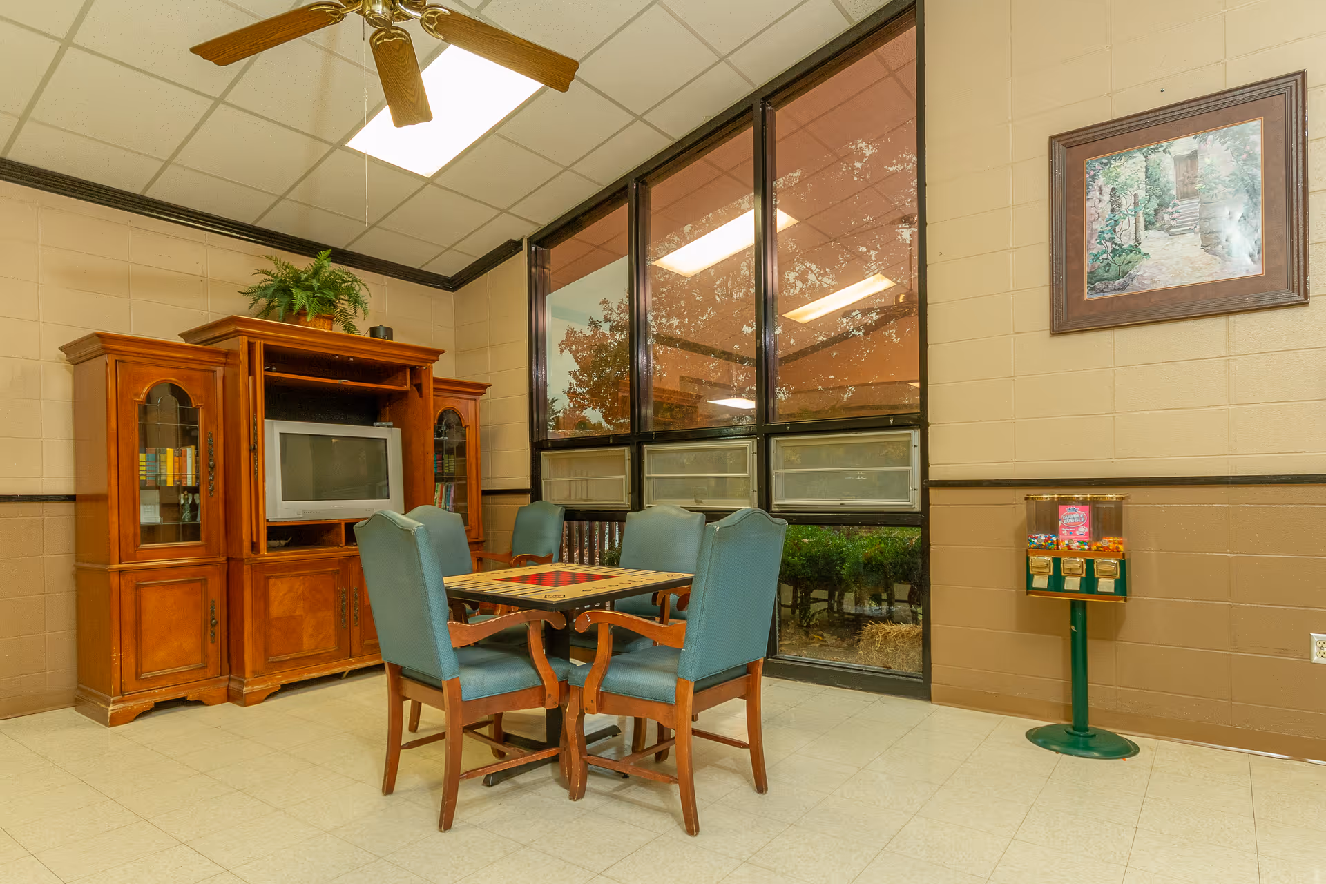 A cozy common area with a wooden entertainment center holding a television and books, a square table with a checkerboard pattern on top surrounded by four blue upholstered chairs, large windows showing outdoor greenery, a ceiling fan, a framed picture on the wall, and a candy vending machine in the corner.