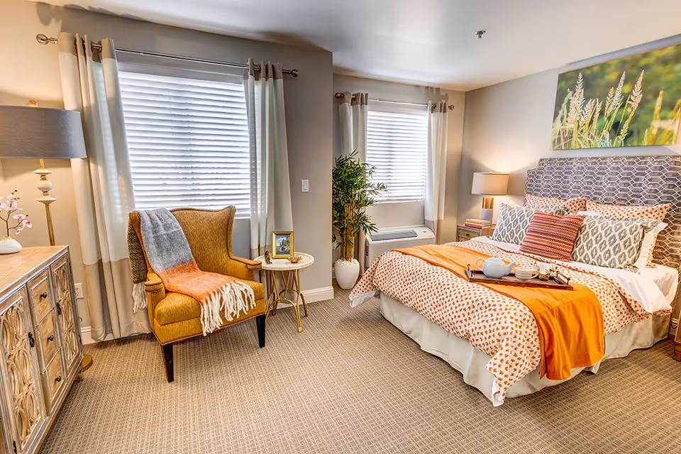 A cozy bedroom in an assisted living facility featuring a bed with patterned bedding and multiple pillows, a tray with a teapot and cups on the bed, a yellow armchair with a colorful throw blanket, a small round side table with a framed photo, a wooden dresser with a lamp and decorative items, two windows with white curtains, a potted plant, and a nature-themed painting on the wall.