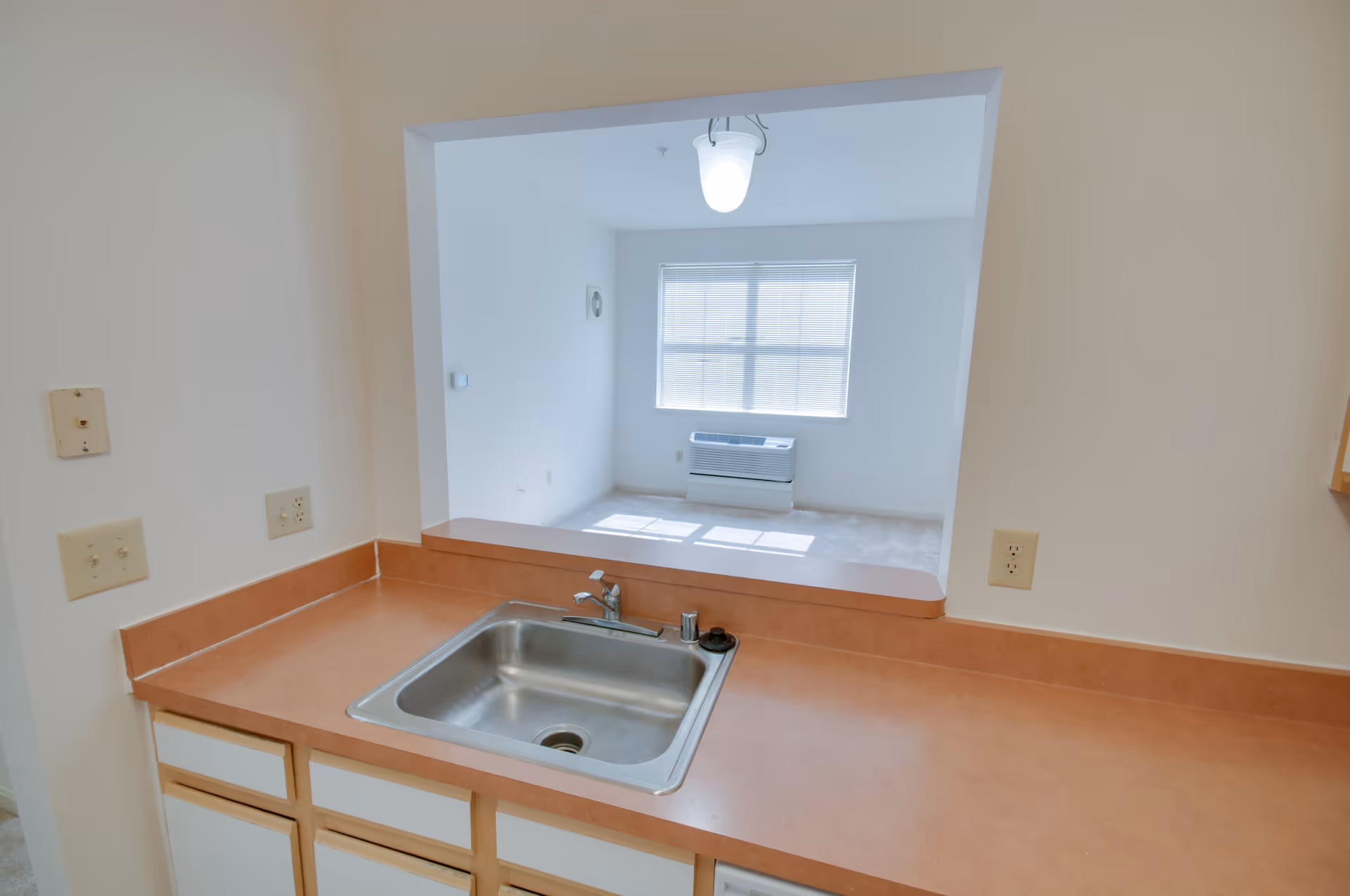View of a kitchen sink with a brown countertop and white cabinets below. There is a pass-through window opening to a bright room with a window, blinds, and a wall-mounted air conditioning unit.
