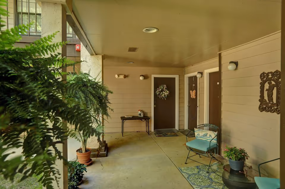 Covered outdoor hallway area with three brown doors, each decorated with a wreath or hanging decoration. There are potted plants, a small table with decorative items, and two cushioned chairs on a patterned rug. The area is shaded with a ceiling and wall-mounted lights.