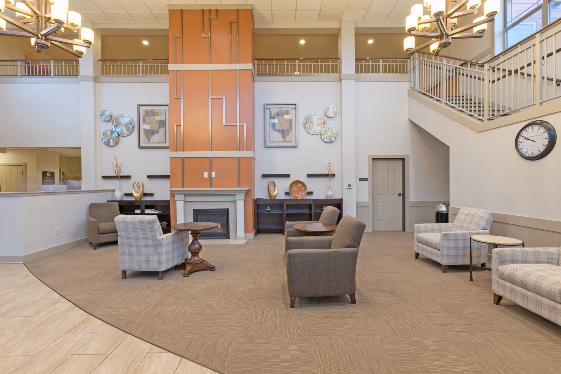 A spacious and well-lit senior living facility common area with a high ceiling, featuring a central fireplace with an orange and beige decorative panel. The room has several comfortable armchairs and small wooden tables arranged around the fireplace. There are decorative wall art pieces and vases on shelves flanking the fireplace. A staircase with a white railing is visible on the right side, along with a wall clock. The flooring is a combination of beige carpet and tile.