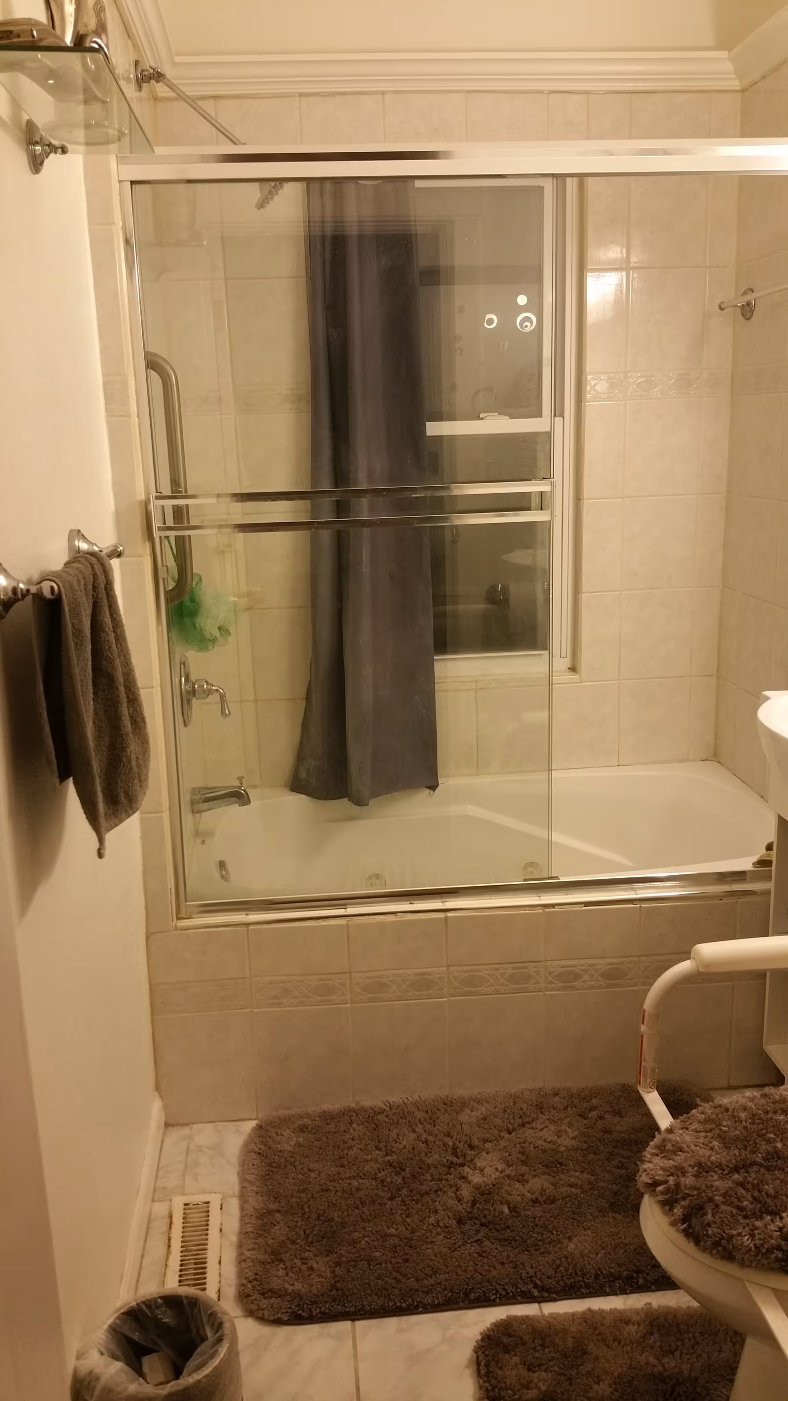 Tiled bathroom with a bathtub and sliding glass shower doors, gray shower curtain, gray bath mats, towel, and a toilet with an armrest.
