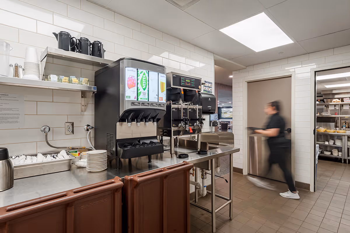 A beverage station in a commercial kitchen area with a soda dispenser, coffee machines, cups, and plates on stainless steel counters. A person in black clothing is walking past a doorway leading to a storage area with shelves holding dishes and food items.