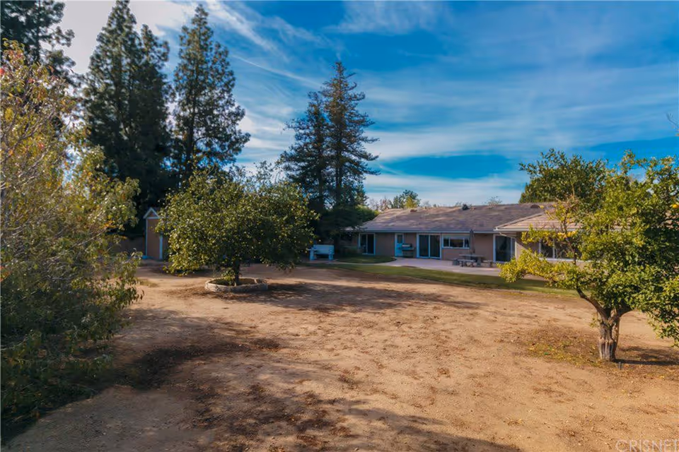 Outdoor view of a senior living facility with a dirt yard, several trees, and a single-story building in the background under a partly cloudy blue sky.