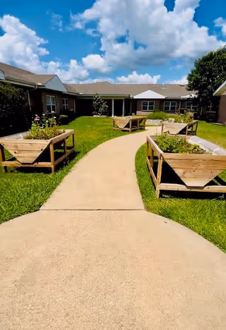 A paved walkway curves through a grassy courtyard with raised wooden garden beds on either side. The courtyard is surrounded by single-story buildings under a partly cloudy blue sky.