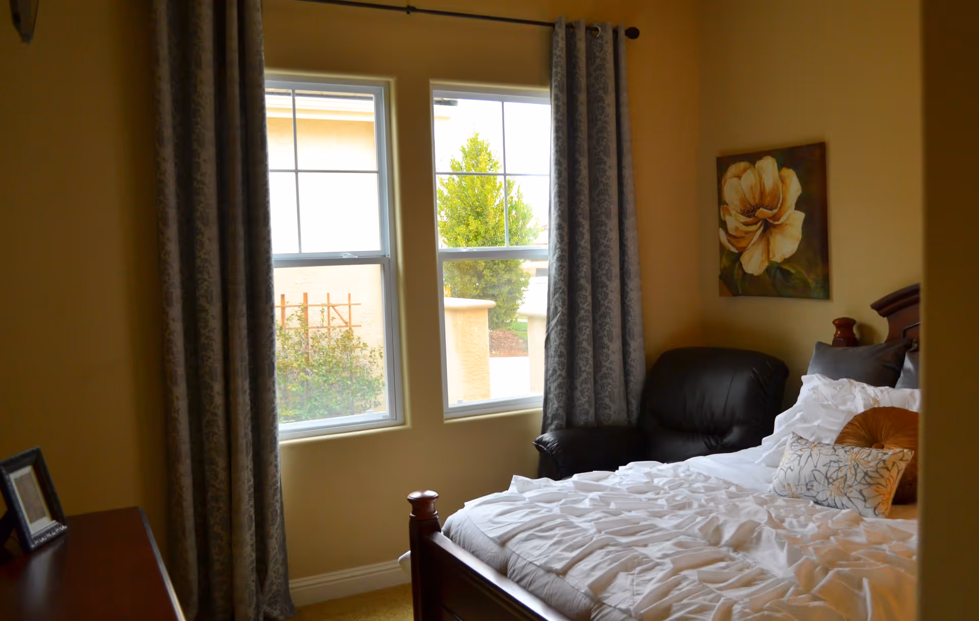 A cozy bedroom with a wooden bed frame, white bedding, and decorative pillows. There is a black armchair in the corner next to two windows with patterned curtains. A floral painting hangs on the wall above the armchair.