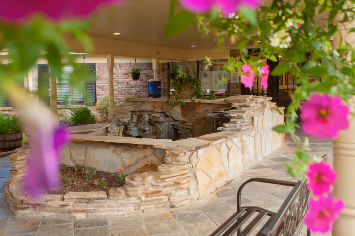 Covered courtyard featuring a tiered stone water feature surrounded by flowering hanging plants and a bench.