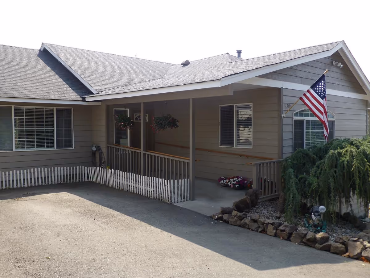 Front exterior of a single-story house with a covered porch and ramp, an American flag, small picket fence, and landscaping.