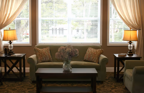 Sunlit sitting area with a sofa, coffee table, side tables with lamps, and large windows with curtains.
