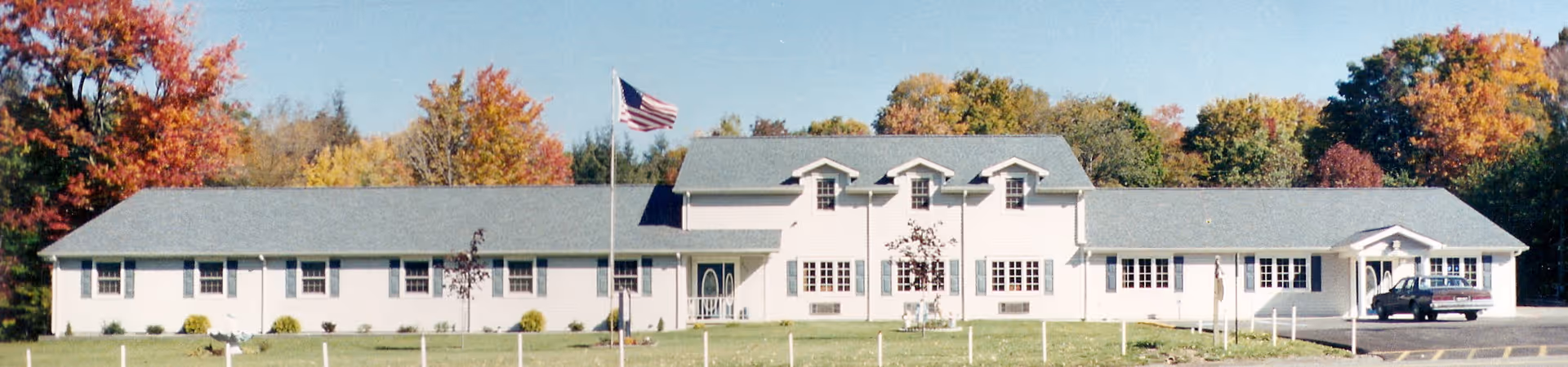 Front exterior view of a long, single-story building with a gray roof and white walls, surrounded by a grassy lawn and trees with autumn foliage. An American flag is flying on a flagpole in front of the building, and a car is parked near the right side.