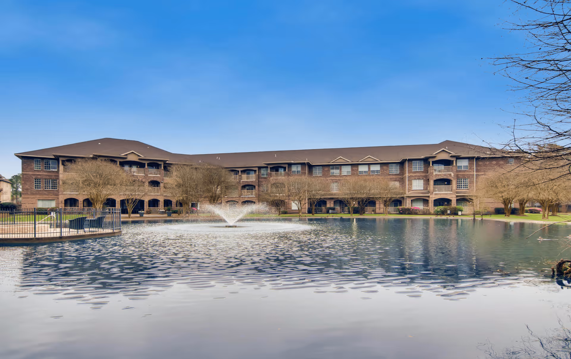 A large three-story brick building with multiple windows and balconies, situated behind a pond with a water fountain in the center. Trees without leaves surround the pond and the building under a clear blue sky.