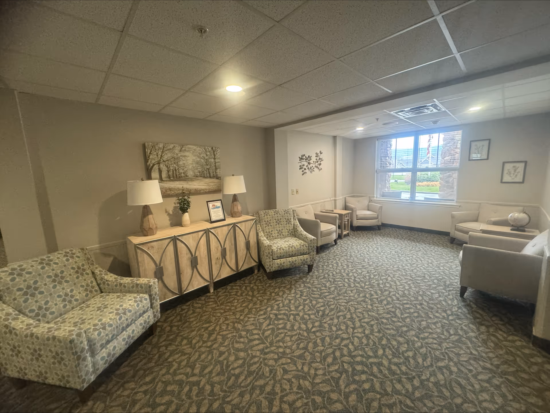 Furnished communal sitting room with multiple armchairs, side tables, a console with lamps and artwork, and a window overlooking the grounds.