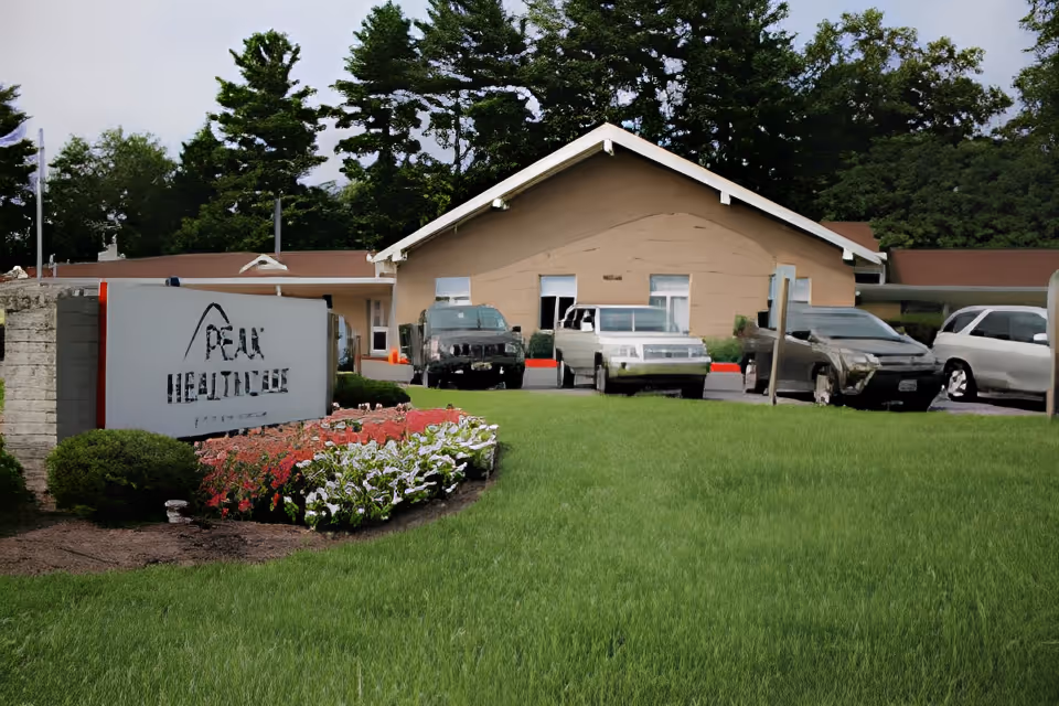 Exterior view of a healthcare facility with a beige building, several parked cars, a green lawn, and a flower bed in front of a sign that reads 'Peak Healthcare'. Trees are visible in the background under a cloudy sky.