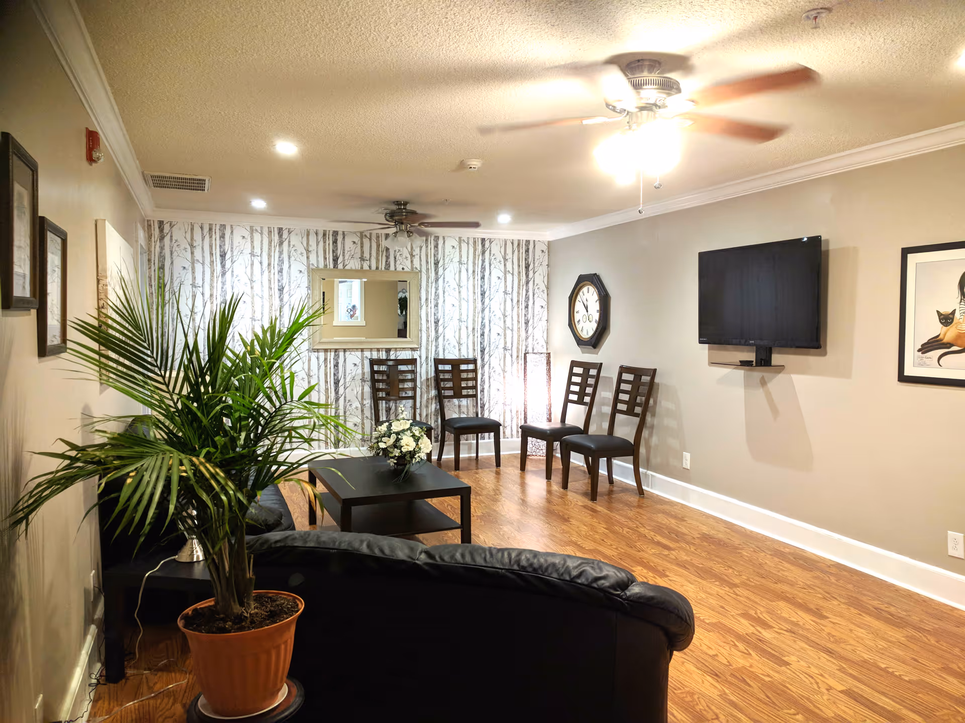 A cozy living room area with wooden flooring, a black leather couch, a coffee table with a flower arrangement, four wooden chairs against a wall with tree-patterned wallpaper, a wall-mounted TV, a large clock, framed artwork, and a potted plant in the foreground.