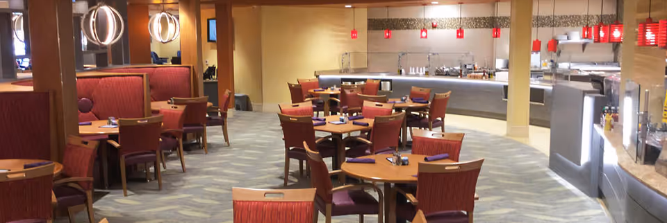 Interior view of a dining area with multiple round wooden tables and red cushioned chairs. There are booths with red upholstery along the left side and a buffet counter with red pendant lights hanging above it on the right side. The floor is carpeted with a patterned design.
