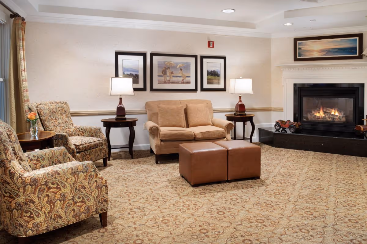 A cozy living room with a beige loveseat, two patterned armchairs, two brown ottomans, and two side tables with lamps. There is a fireplace with a fire burning and a framed picture above it. The walls have three framed pictures and the floor is carpeted with a patterned design.