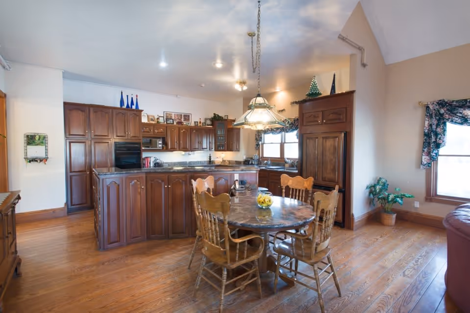 A spacious kitchen and dining area with wooden cabinets and hardwood floors. The kitchen features a large island with a marble countertop, built-in oven, microwave, and a refrigerator with wooden paneling. A round dining table with four wooden chairs is positioned in front of the island, with a hanging light fixture above it. There are windows with floral curtains and a potted plant near the corner.