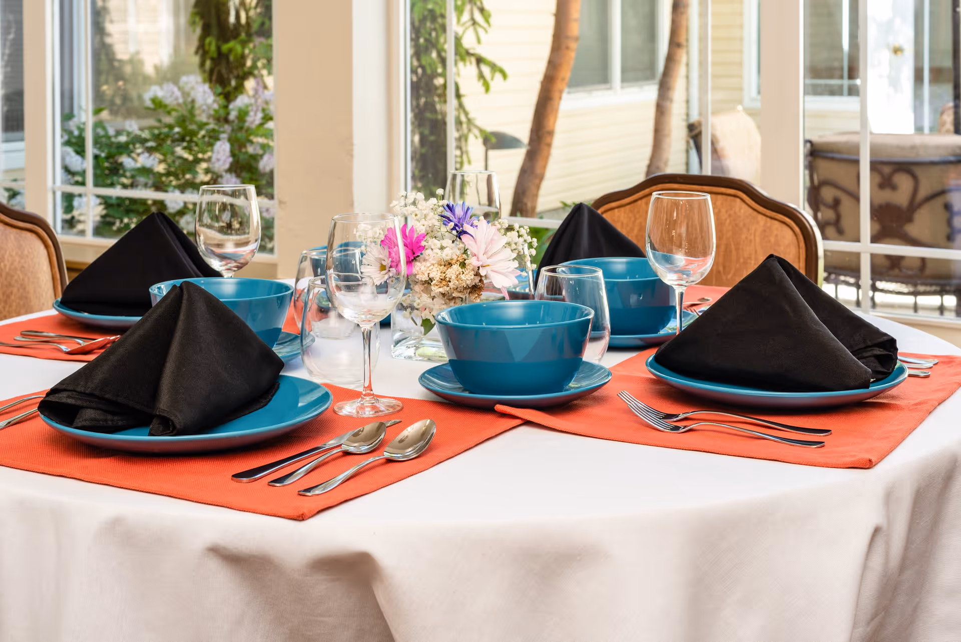 A dining table set for four with teal bowls and plates, black folded napkins, silverware, and clear wine glasses on an orange placemat. A small floral centerpiece with pink, purple, and white flowers is in the middle. The table is near large windows showing outdoor greenery.