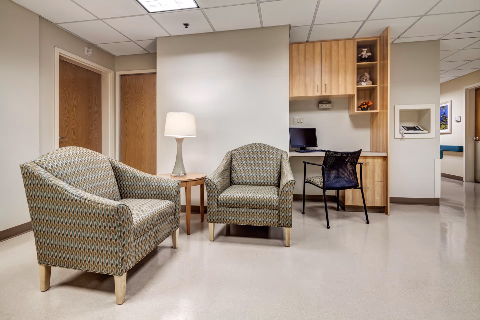 A small seating area in a healthcare facility with two patterned armchairs and a round wooden side table with a lamp. To the right, there is a built-in wooden desk with a computer monitor, a black mesh chair, and shelves with decorative items. The walls are light-colored, and there are two closed wooden doors in the background.