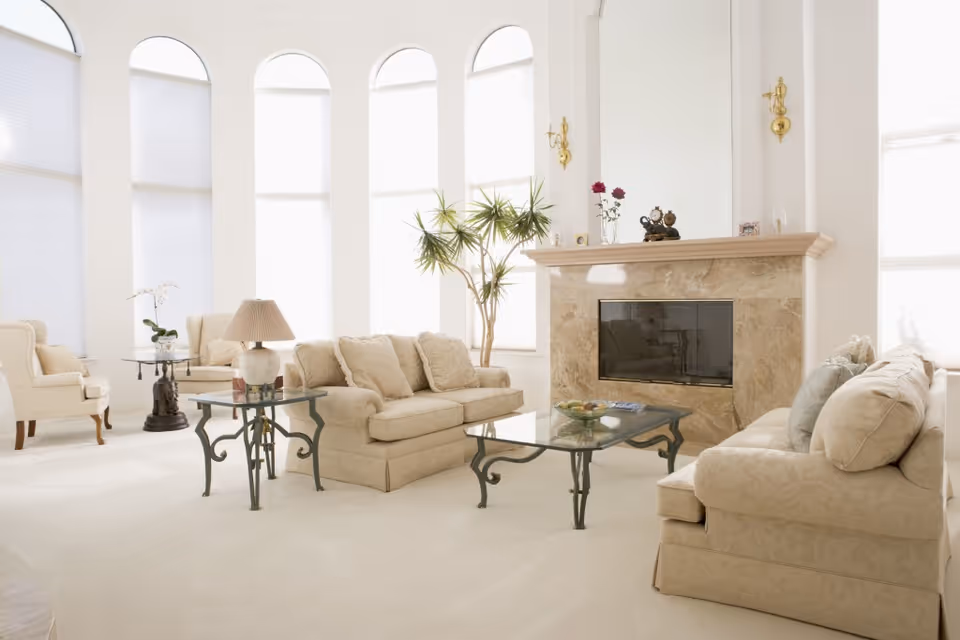 Bright airy living room with beige sofas, glass coffee tables, and a marble fireplace beneath tall arched windows.