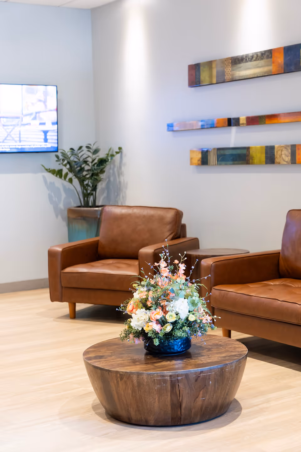 A cozy seating area with two brown leather armchairs and a round wooden coffee table in the center holding a floral arrangement. There is a potted plant in the corner and colorful abstract wall art on the white wall behind the chairs.