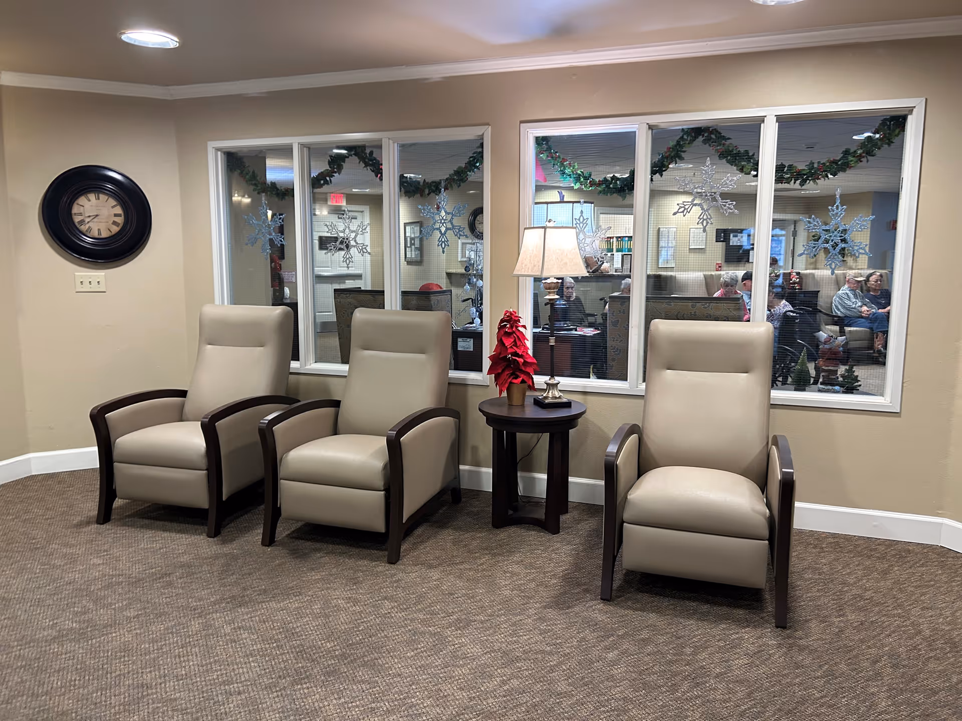 A cozy seating area with three beige armchairs arranged around a small round wooden table with a lamp and a red poinsettia plant. Behind the chairs is a large window decorated with snowflake decals and garlands, through which a room with several elderly people sitting on couches is visible. A round wall clock is mounted on the beige wall to the left.