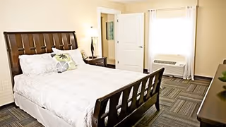 A bright bedroom with a wooden bed frame, white bedding, decorative pillows, a window with white curtains, a door, and a dresser with a plant on top.