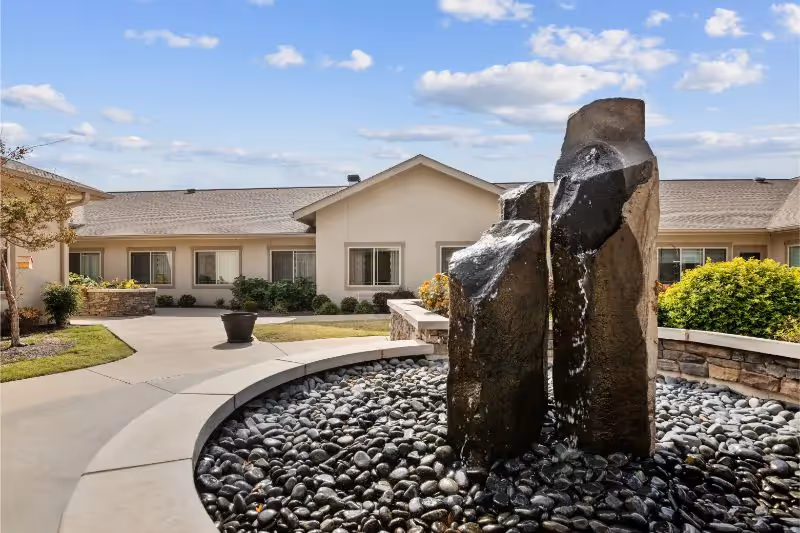A stone water fountain set in a circular pebble bed in the courtyard of a single-story senior living building under a blue sky.