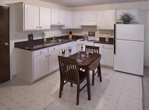 A clean and well-lit kitchen featuring white cabinets, a white refrigerator, a stove with a kettle, and a dark countertop. In the center, there is a small wooden dining table with two chairs, set with glasses and napkins. The floor has a light-colored patterned linoleum, and there is a wooden door on the left side of the image.