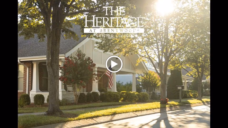 Exterior view of The Heritage at Brentwood senior living facility showing a row of single-story buildings with porches, surrounded by trees and shrubs in sunlight.